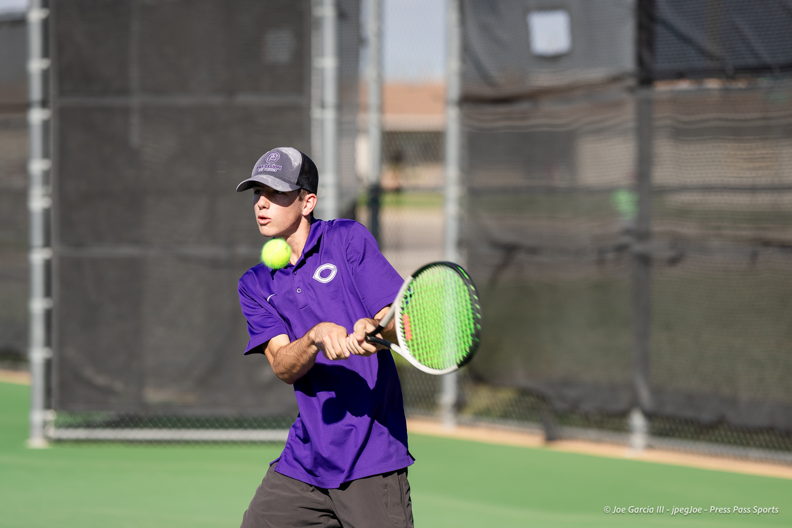 canyon-regional-tennis-26 | Press Pass Sports Male tennis player in a purple polo swings at a tennis ball on an outdoor court, concentrating on the shot with a green racket.