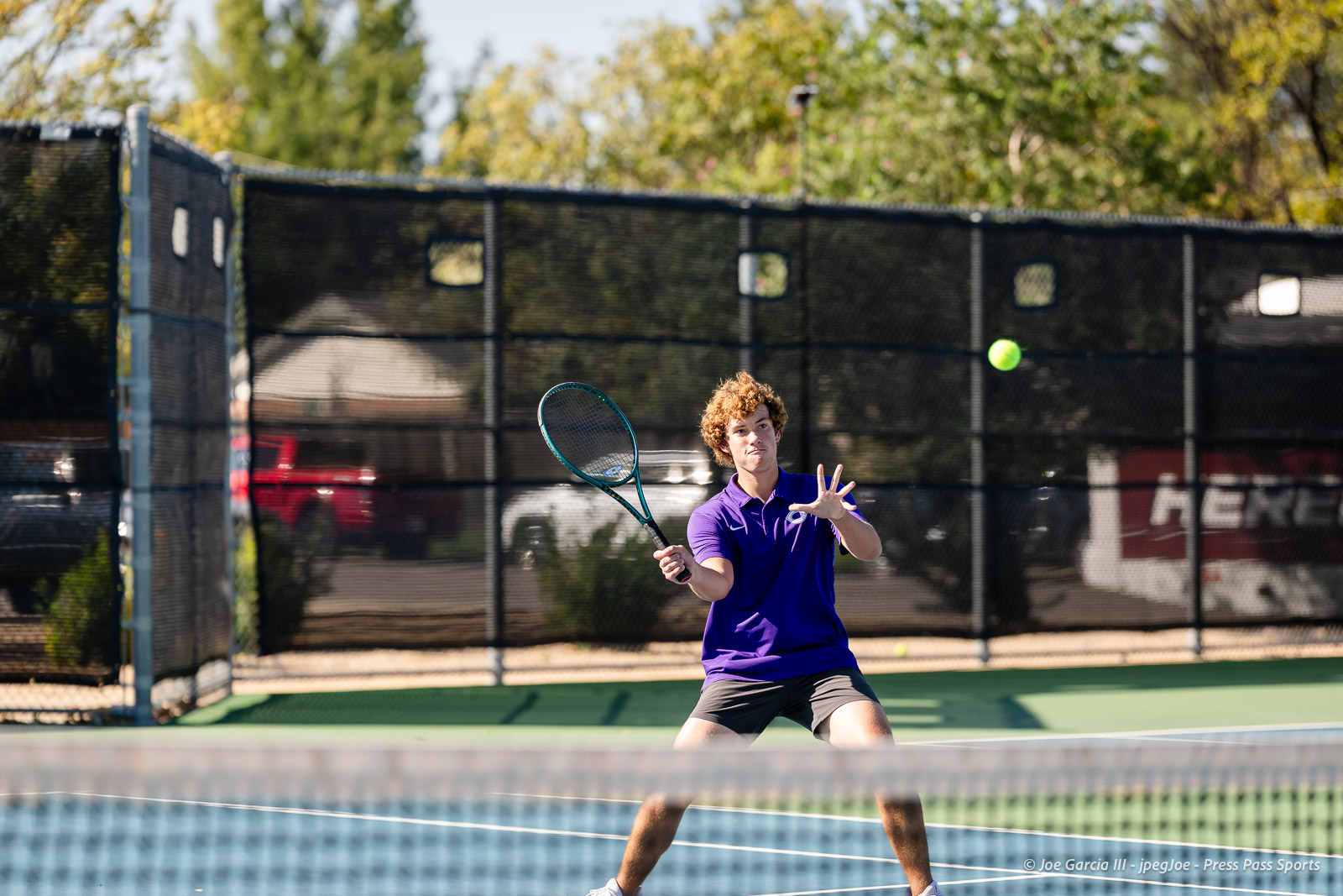 canyon-regional-tennis-27 | Press Pass Sports Young male tennis player wearing a purple polo swings his racket on a blue hardcourt, ball in midair near the fence behind him.