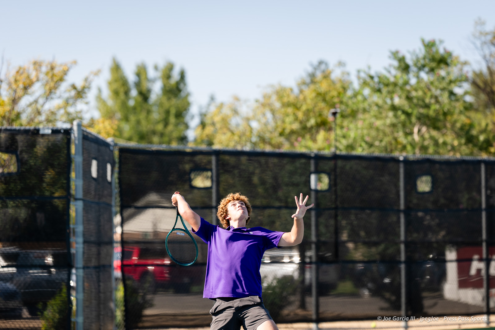 canyon-regional-tennis-29 | Press Pass Sports Tennis player in a purple shirt serving a ball on an outdoor court with a chain-link fence behind him.