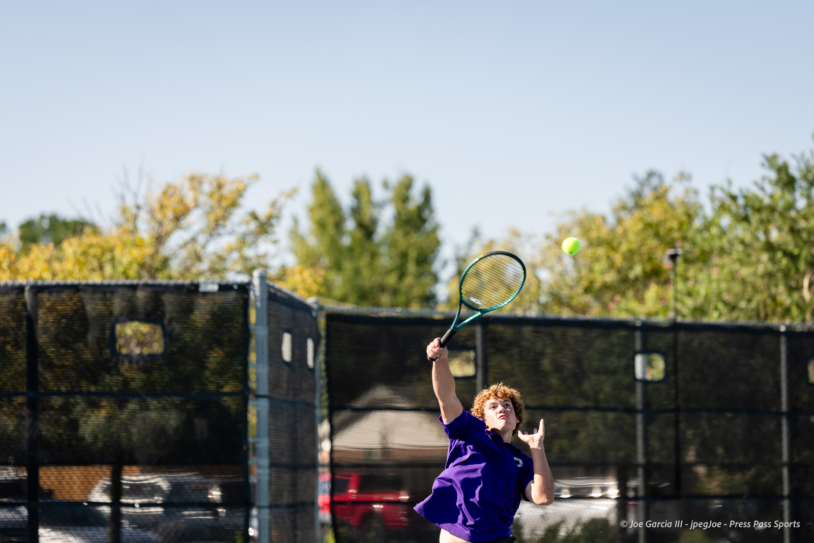 canyon-regional-tennis-31 | Press Pass Sports Young tennis player in a purple shirt reaches up with a teal racket to hit a bright yellow ball on an outdoor court behind a chain-link fence when the sun is out.