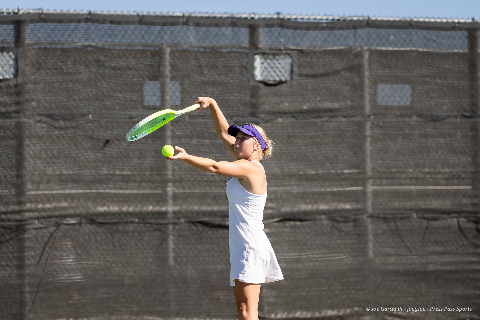 canyon-regional-tennis-32 | Press Pass Sports Female tennis player in a white dress serves the ball on an outdoor court, wearing a purple visor.
