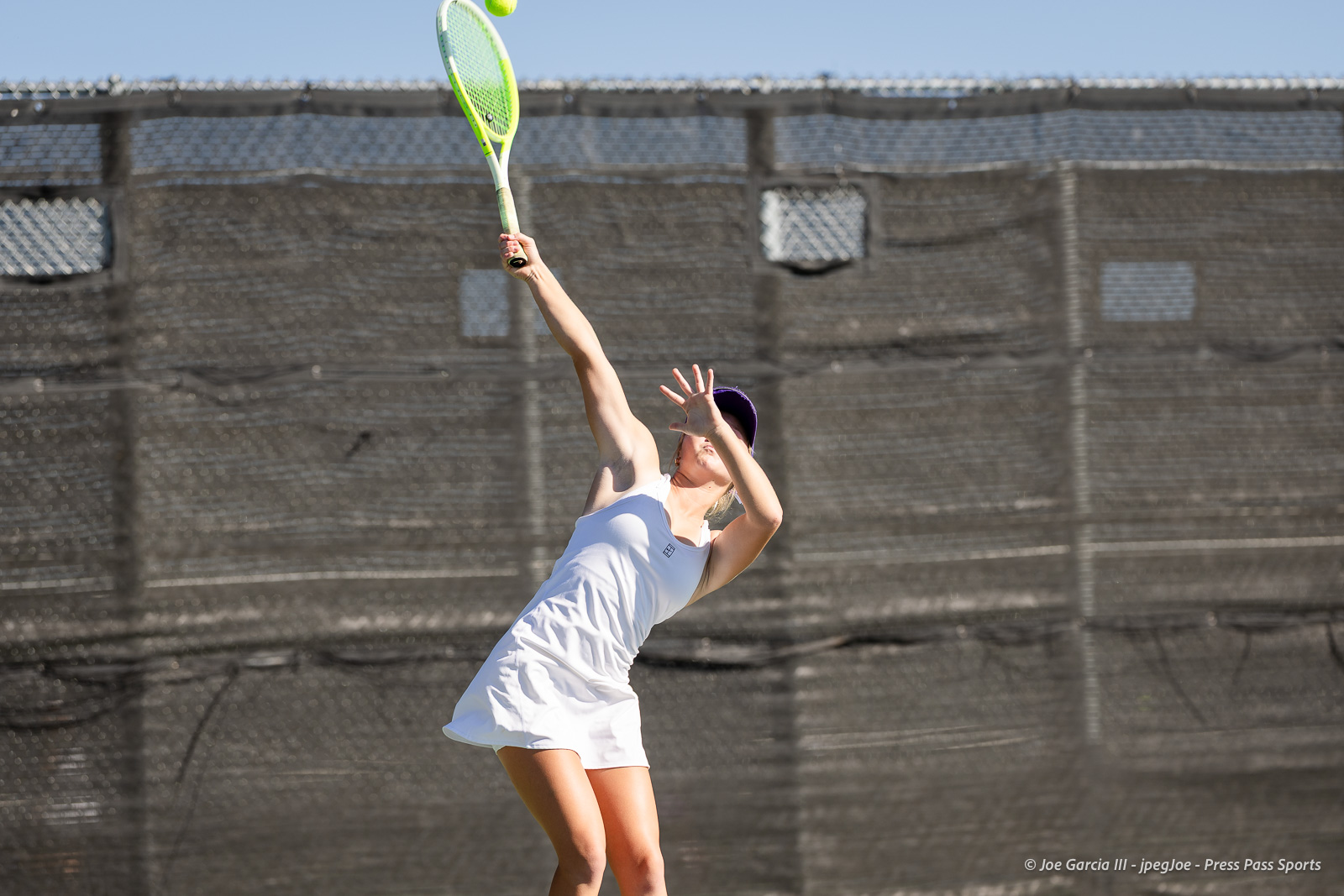 canyon-regional-tennis-33 | Press Pass Sports Female tennis player in a white outfit serves the ball on an outdoor court with a tall black mesh fence behind her.