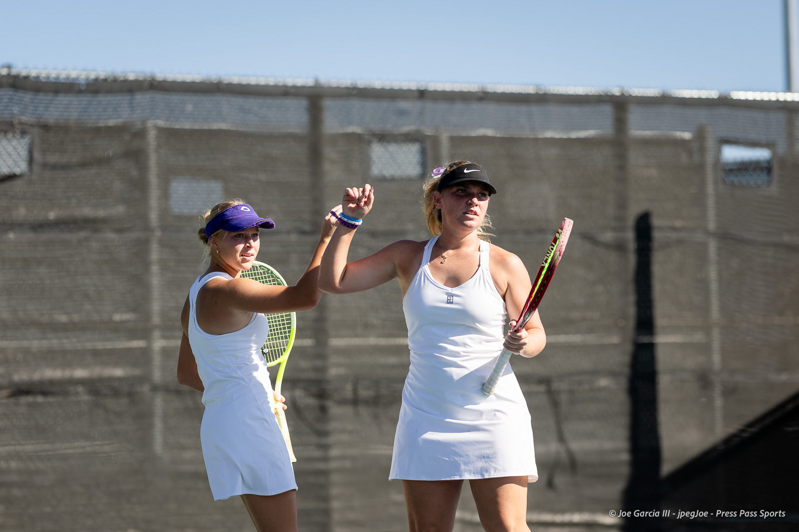 canyon-regional-tennis-35 | Press Pass Sports Two female tennis players in white outfits high-fiving on a sunlit court with rackets in hand.