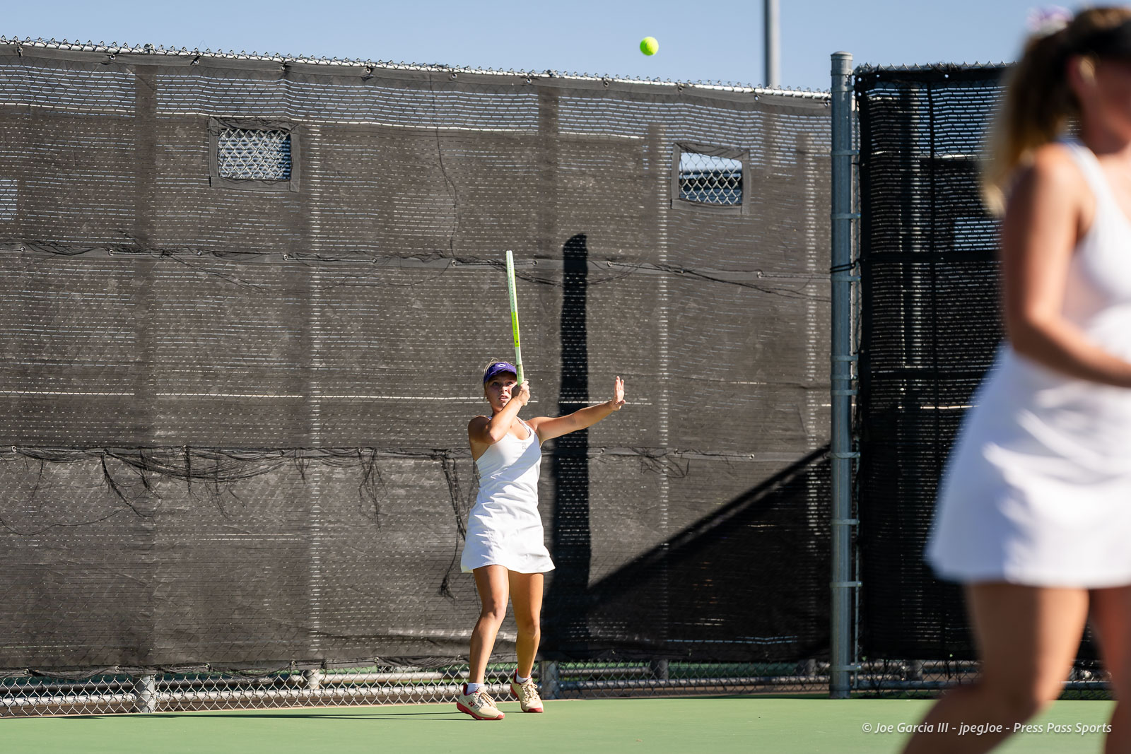 canyon-regional-tennis-38 | Press Pass Sports Female tennis player in a white outfit prepares to hit a backhand on an outdoor court, with a bright ball in the air behind her.