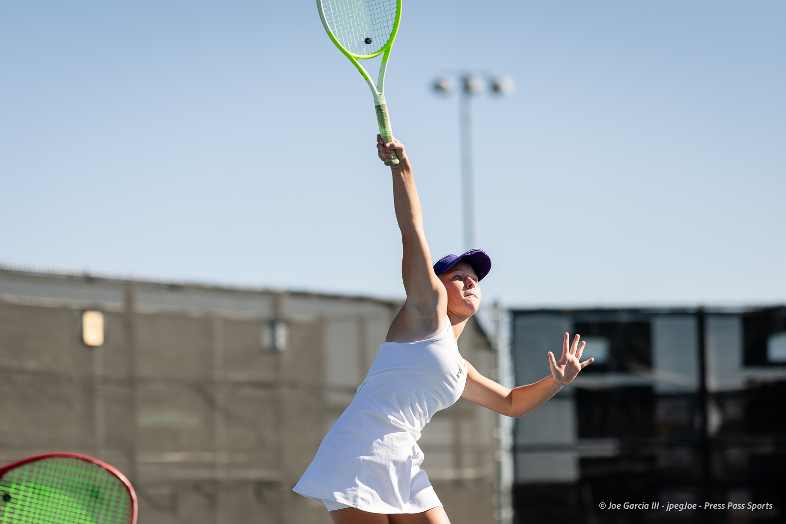 canyon-regional-tennis-41 | Press Pass Sports Female tennis player in a white dress serves the ball on an outdoor court under a clear blue sky, wearing a purple cap.