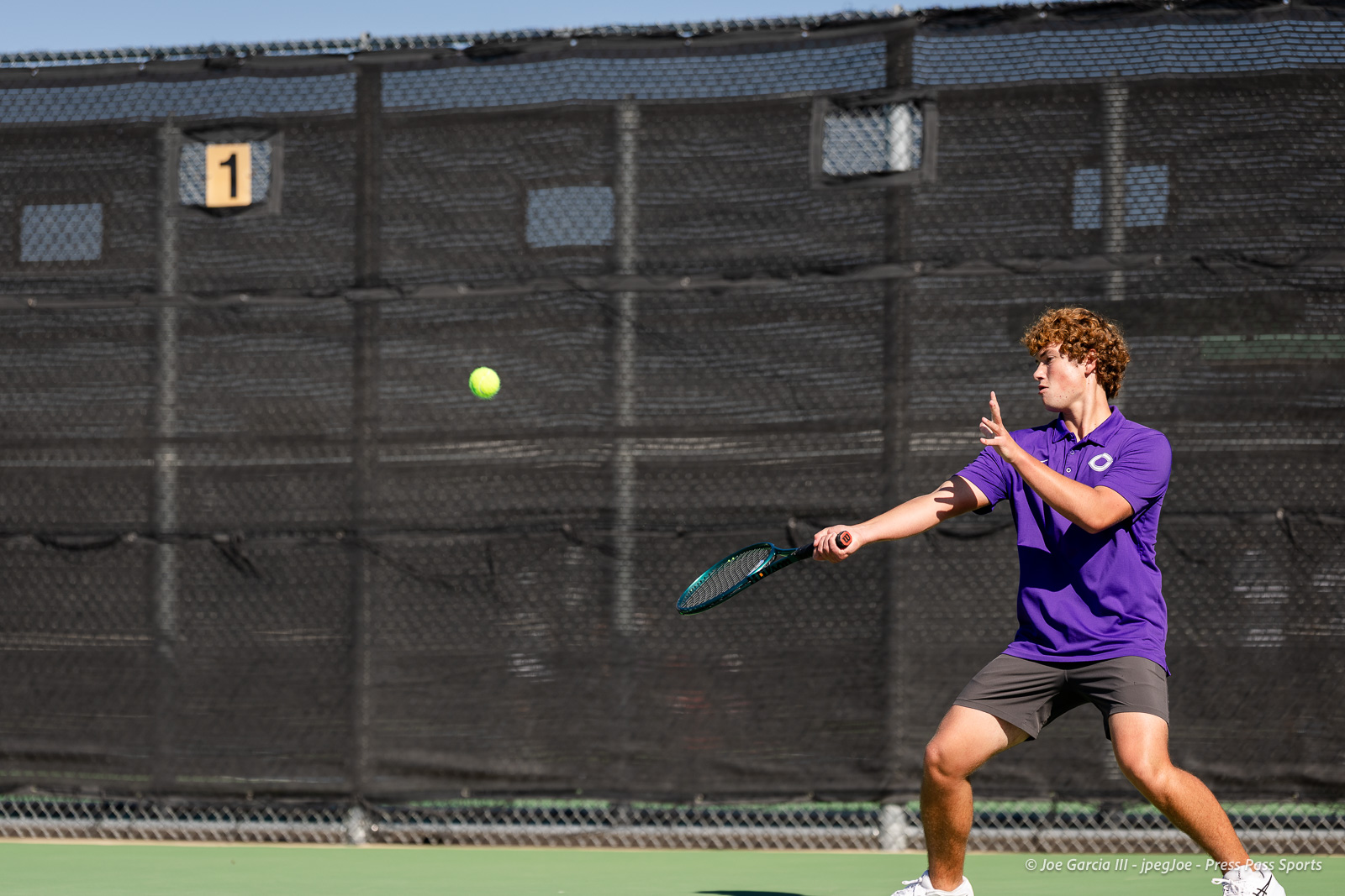 canyon-regional-tennis-43 | Press Pass Sports Tennis player in a purple polo swings a racket at a neon yellow ball on an outdoor court with a chain-link fence behind him.