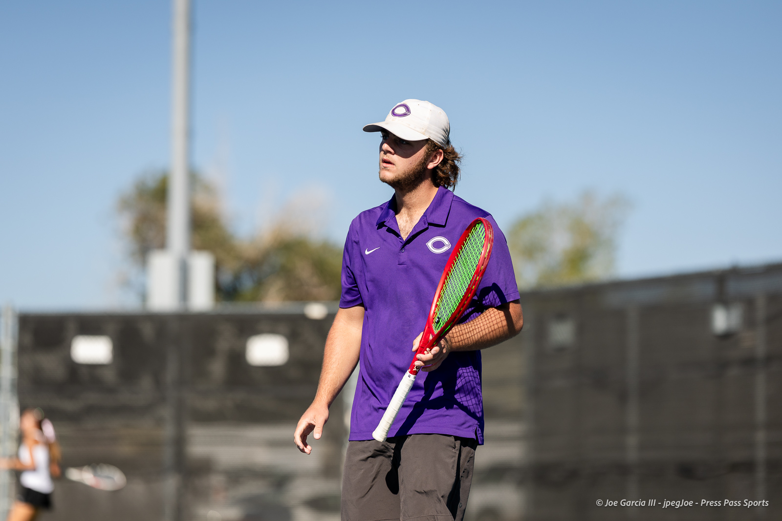 canyon-regional-tennis-45 | Press Pass Sports Male tennis player in a purple polo and white cap stands on an outdoor court holding a red-and-green racket, looking to the side.