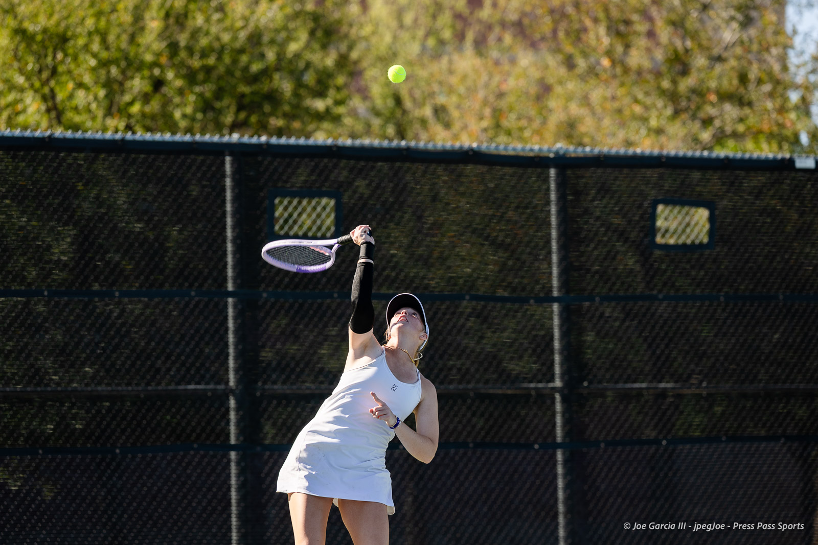 canyon-regional-tennis-46 | Press Pass Sports Female tennis player in a white outfit serves the ball on an outdoor court with a chain-link fence behind her.
