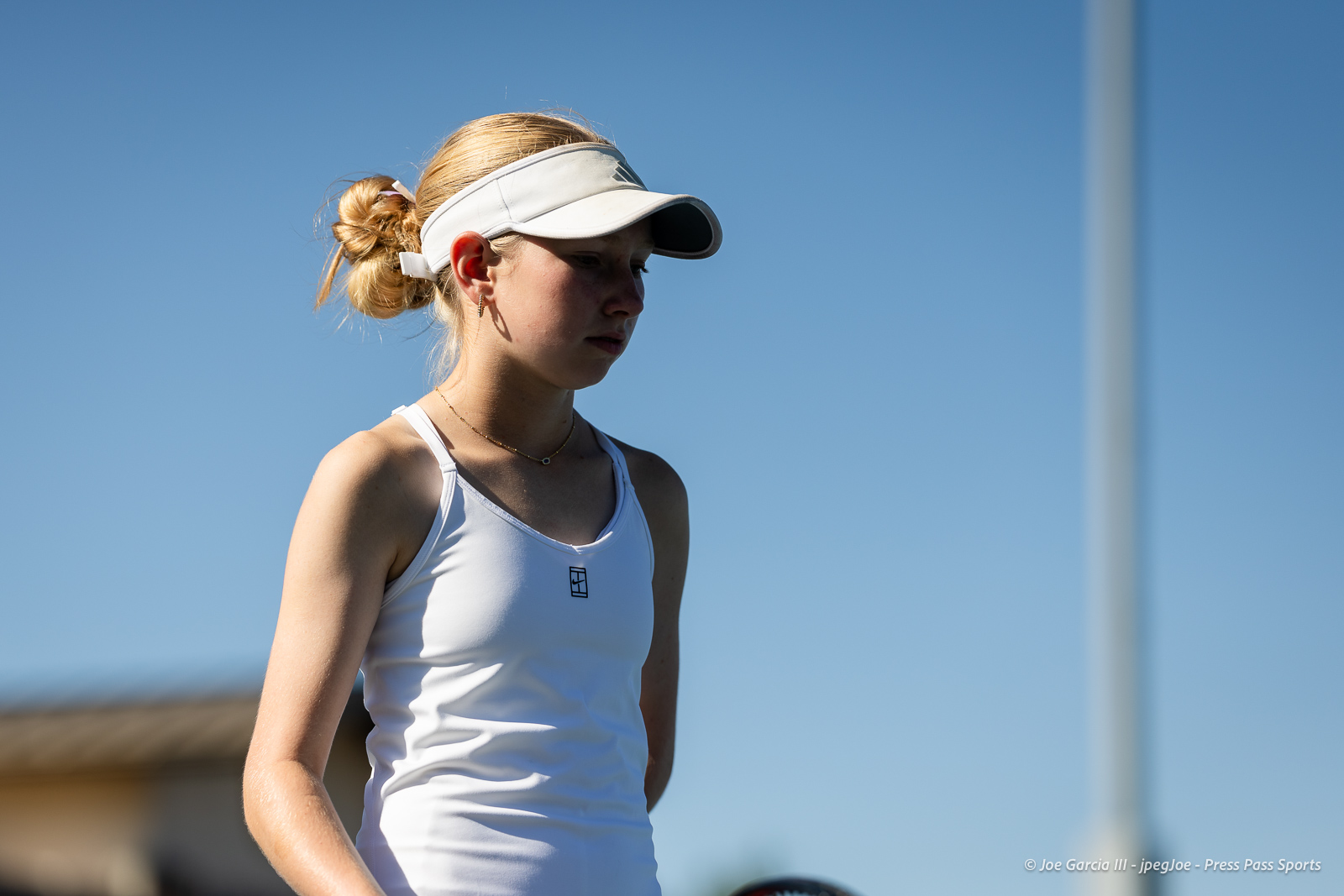 canyon-regional-tennis-48 | Press Pass Sports Young woman in a white tank top and white visor standing outdoors against a clear blue sky, looking downward with a focused expression.