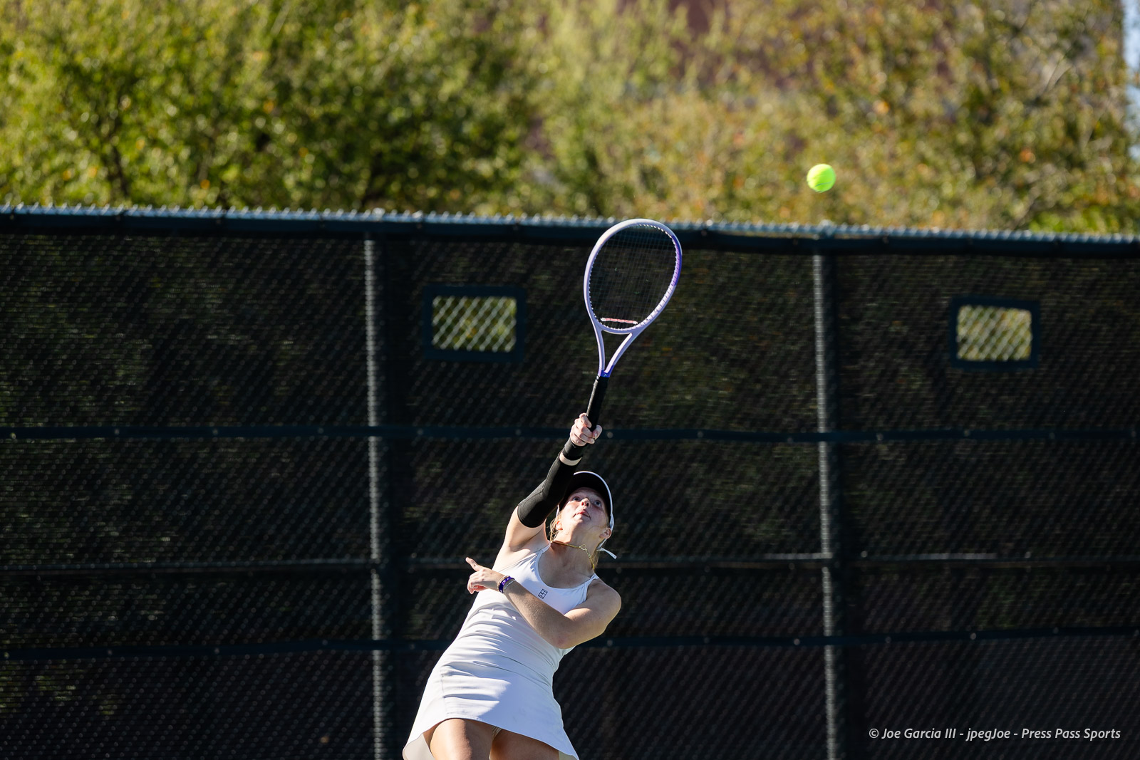 canyon-regional-tennis-49 | Press Pass Sports Female tennis player in a white outfit serves the ball on an outdoor court, racket raised high and ball in midair behind a chain-link fence.