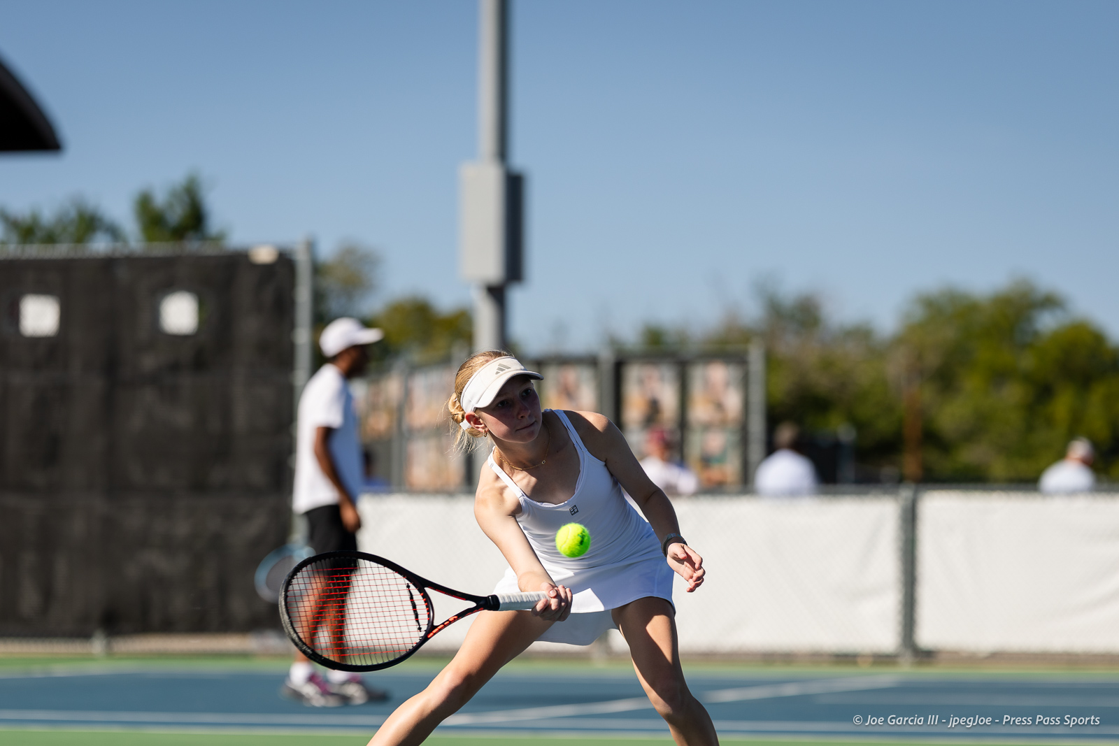 canyon-regional-tennis-50 | Press Pass Sports Female tennis player in a white outfit lunges to hit a bright tennis ball with a black racket on an outdoor court.