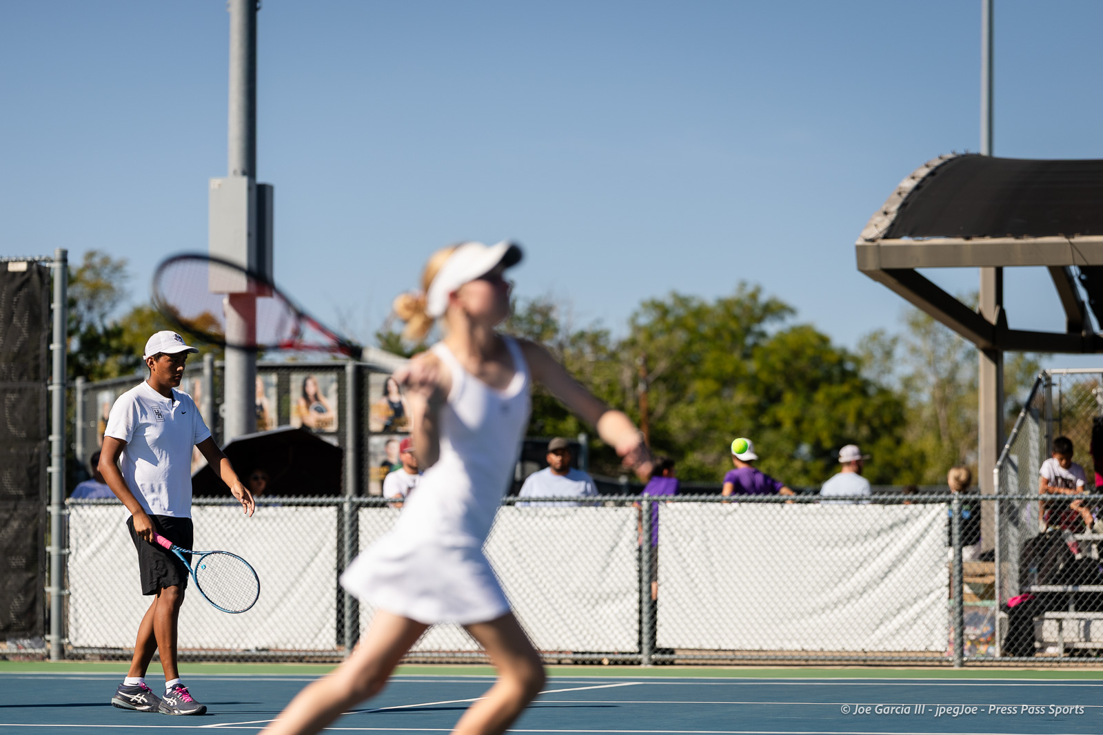 canyon-regional-tennis-52 | Press Pass Sports Tennis players on a blue outdoor court; foreground woman in white swings a racket while a man in a white shirt and cap watches from the left.