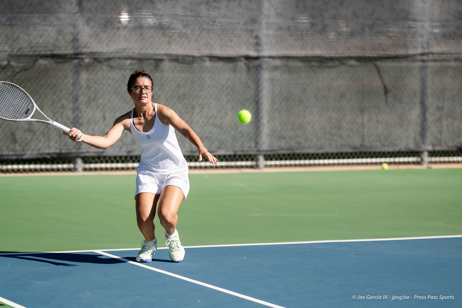 canyon-regional-tennis-55 | Press Pass Sports Female tennis player in white athletic wear lunges to hit a ball with a racket on an outdoor court, focused on the shot as the ball approaches.