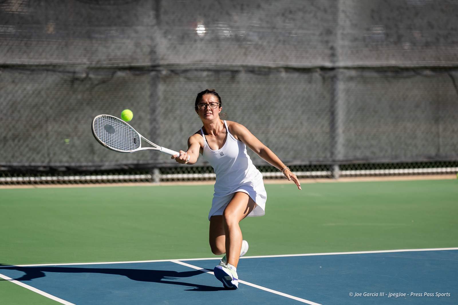 canyon-regional-tennis-56 | Press Pass Sports Female tennis player in a white outfit lunges to hit a bright ball with a racket on a blue court, fence in the background