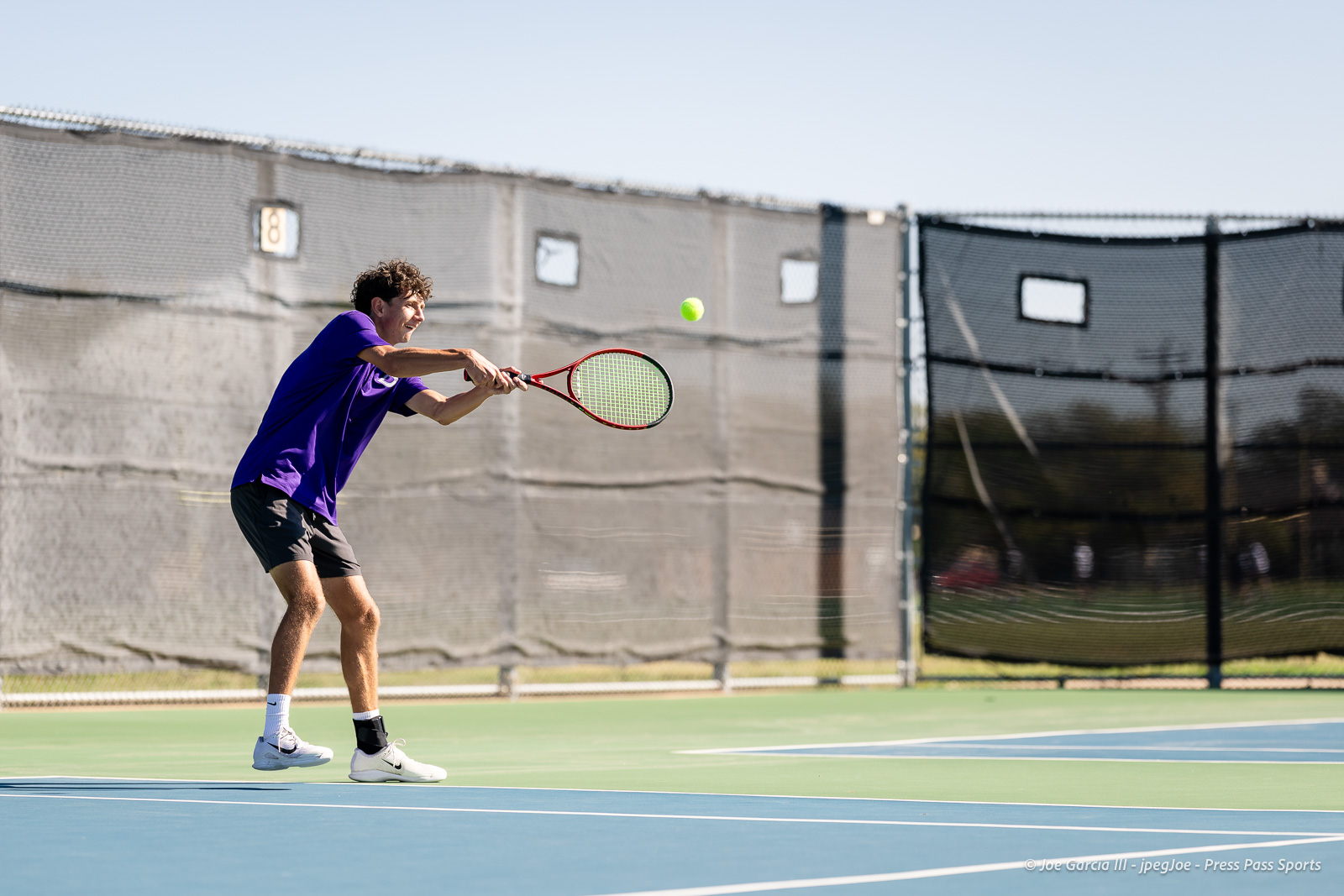 canyon-regional-tennis-57 | Press Pass Sports Tennis player in a purple shirt hitting a forehand on a blue court with a tennis ball mid-air, surrounded by chain-link fencing.