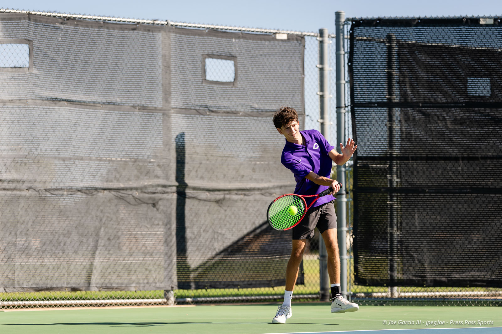 canyon-regional-tennis-58 | Press Pass Sports Male tennis player in a purple shirt hits a forehand on an outdoor court with a green surface and chain-link fences behind him.