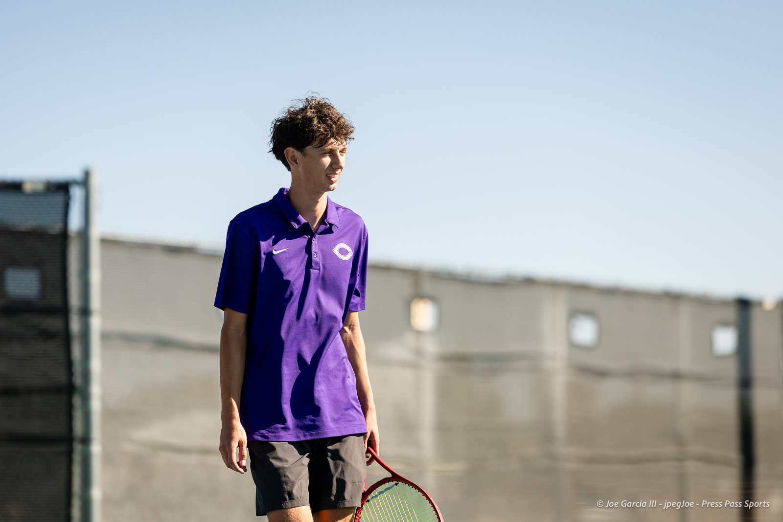 canyon-regional-tennis-59 | Press Pass Sports Young man in a purple polo shirt stands on an outdoor tennis court, holding a red racket as he looks to the side.