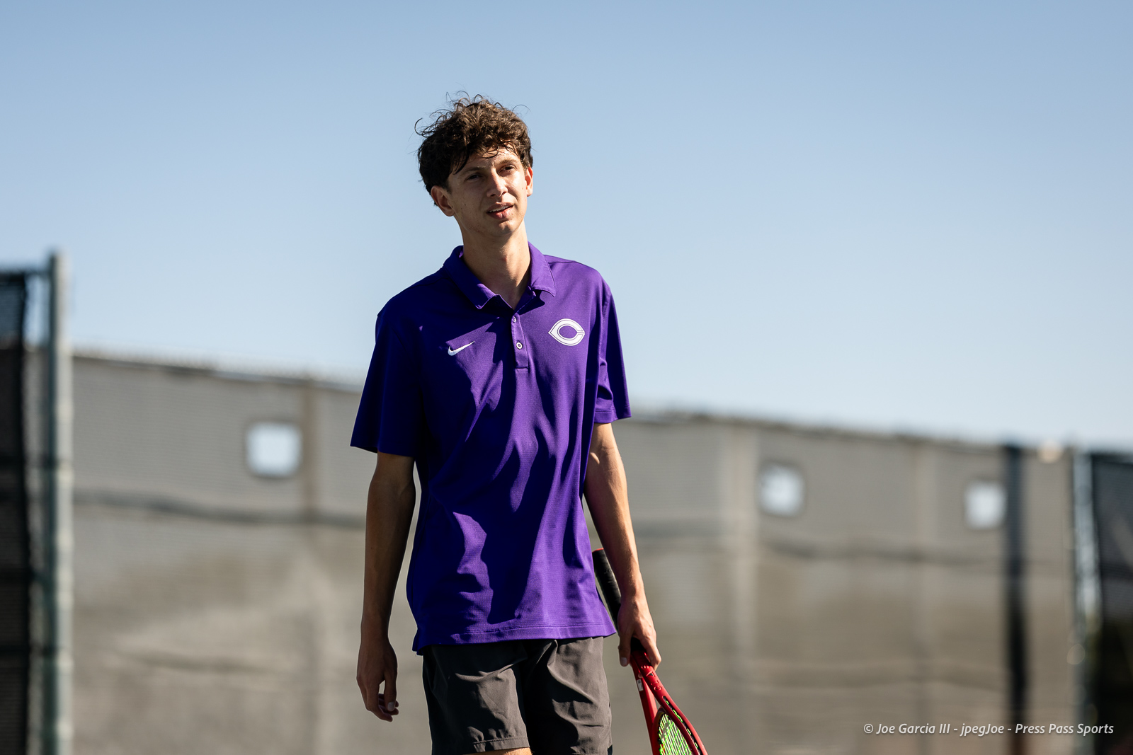 canyon-regional-tennis-60 | Press Pass Sports Young male tennis player in a purple polo with a white logo, holding a red racket on an outdoor court with a fence in the background.