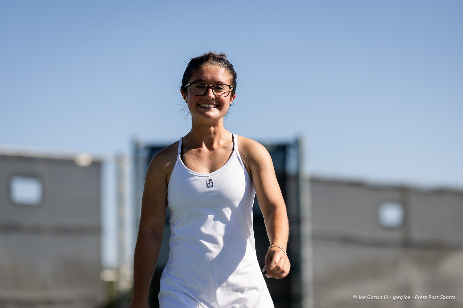 canyon-regional-tennis-62 | Press Pass Sports Smiling woman in a white tank top walks outdoors on a sunny day, wearing glasses and a hair bun.
