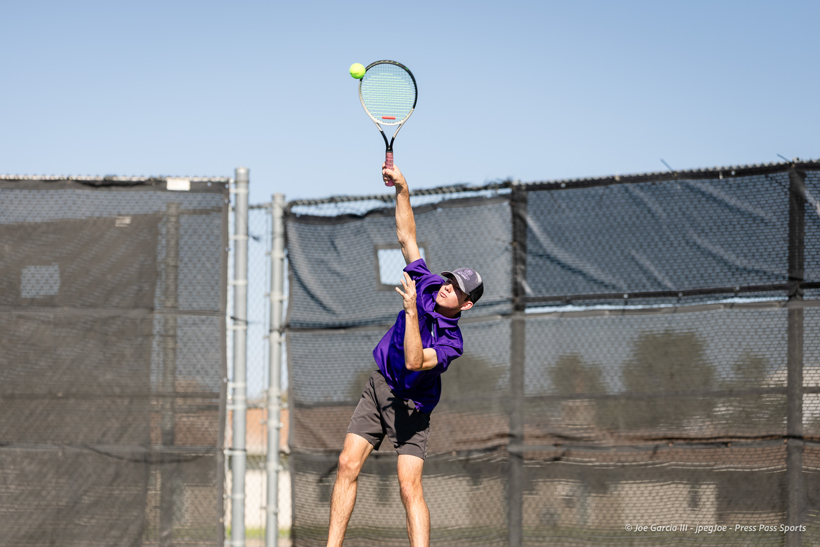 canyon-regional-tennis-64 | Press Pass Sports Male tennis player in a purple shirt and cap mid-serve, racket raised high with a bright tennis ball near the strings on an outdoor court behind a fence.
