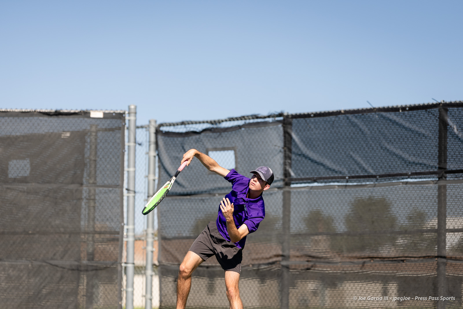 canyon-regional-tennis-65 | Press Pass Sports Male tennis player in a purple shirt and cap swinging a forehand on an outdoor court with a chain-link fence behind him.