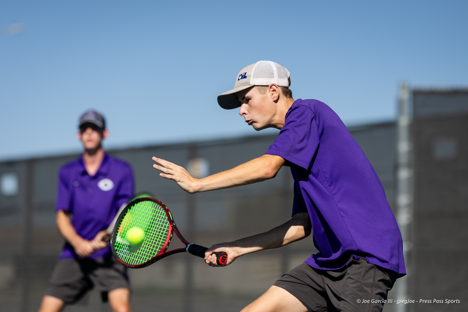 canyon-regional-tennis-66 | Press Pass Sports Tennis player in a purple shirt and gray cap lunges to hit a forehand with a neon-green racket, a ball in midair nearby.
