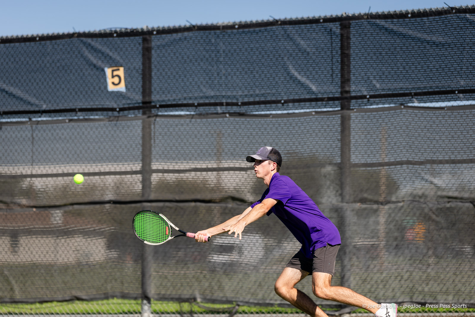 canyon-regional-tennis-67 | Press Pass Sports Tennis player in a purple shirt and cap lunges to hit a forehand on an outdoor court with a chain-link fence behind him.