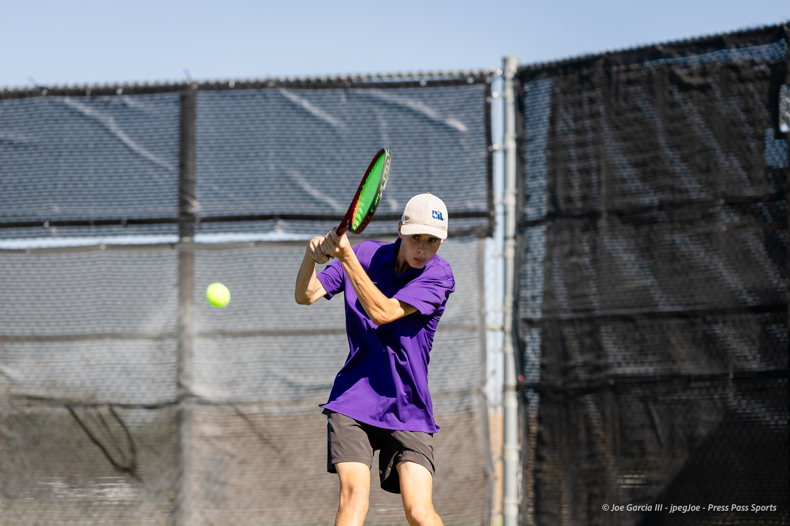 canyon-regional-tennis-68 | Press Pass Sports Tennis player in a purple shirt and beige cap swings at a bright yellow ball on an outdoor court with a chain-link fence behind him.