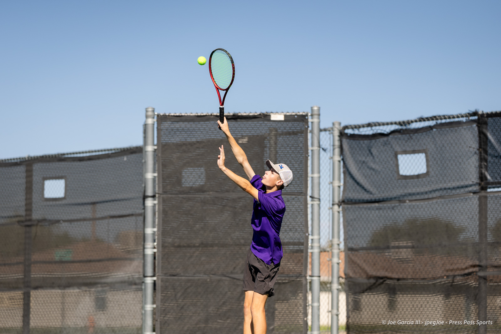 canyon-regional-tennis-69 | Press Pass Sports Male tennis player in a purple shirt and cap serves on an outdoor court, racket raised and ball in midair against a blue sky backdrop.