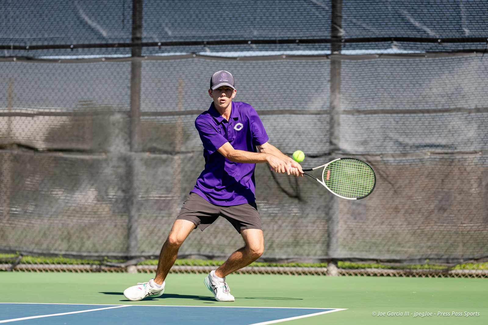 canyon-regional-tennis-70 | Press Pass Sports Male tennis player in a purple polo and gray cap hits a backhand on an outdoor court as a tennis ball approaches.