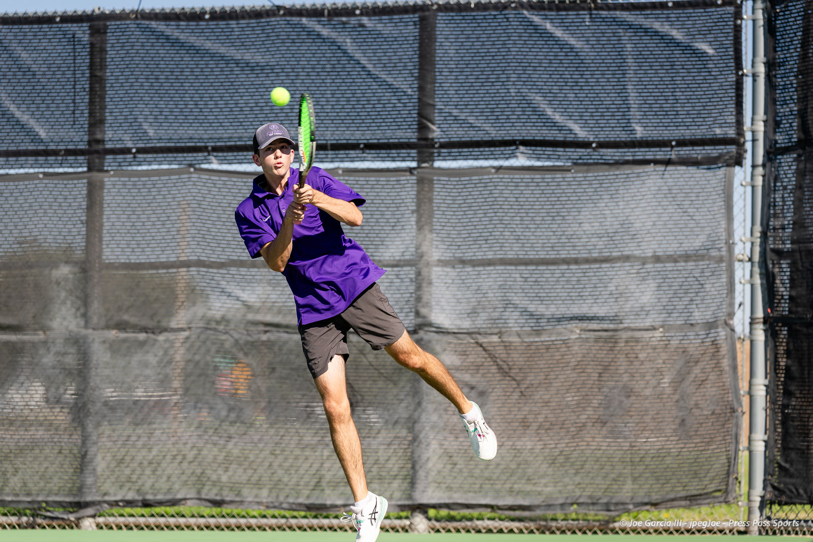 canyon-regional-tennis-73 | Press Pass Sports Male tennis player in a purple shirt and gray shorts leaps to hit a forehand on an outdoor court with a netted fence in the background, ball midair.