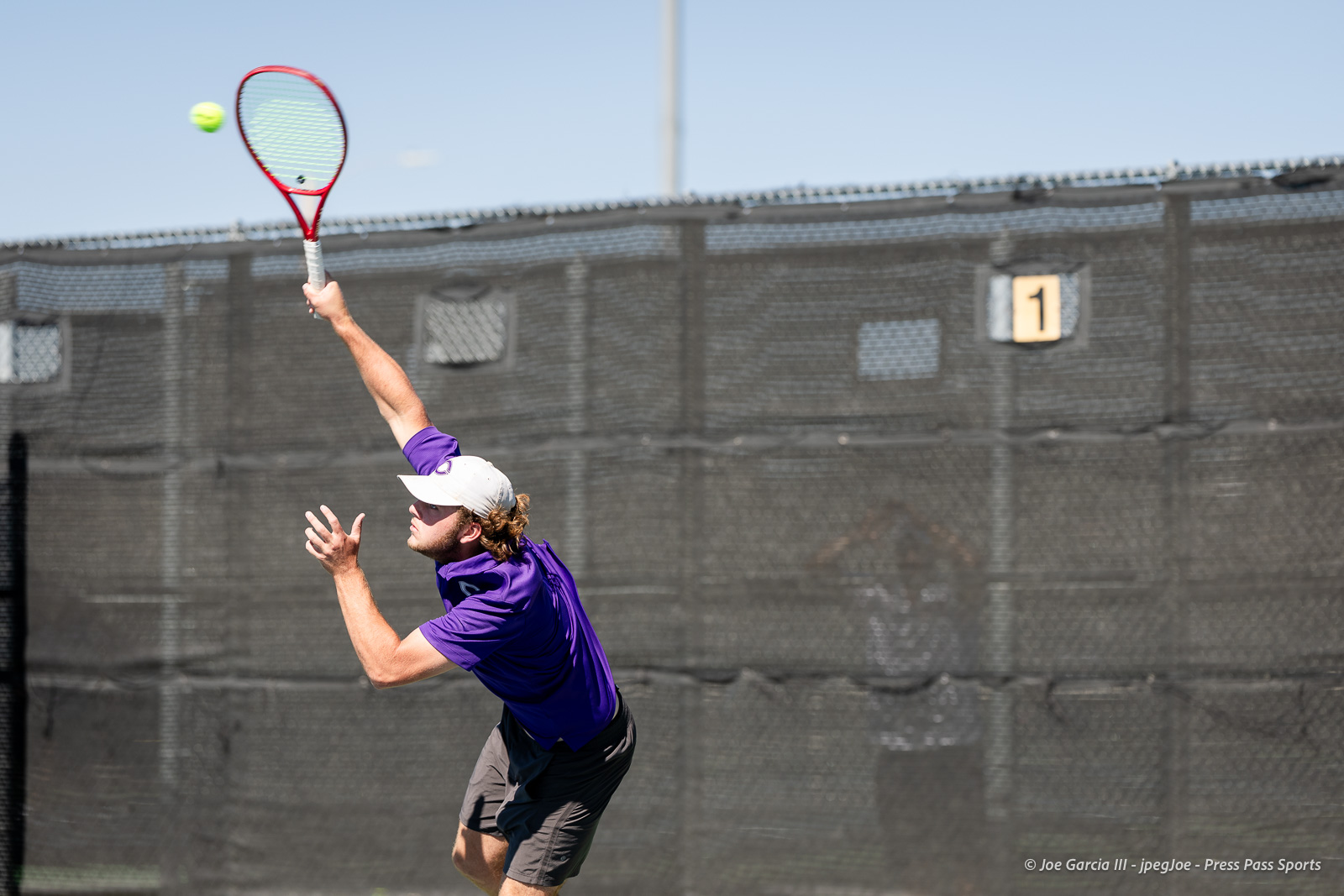 canyon-regional-tennis-74 | Press Pass Sports Tennis player in a purple shirt and white cap reaching to serve on an outdoor court with a chain-link fence behind.
