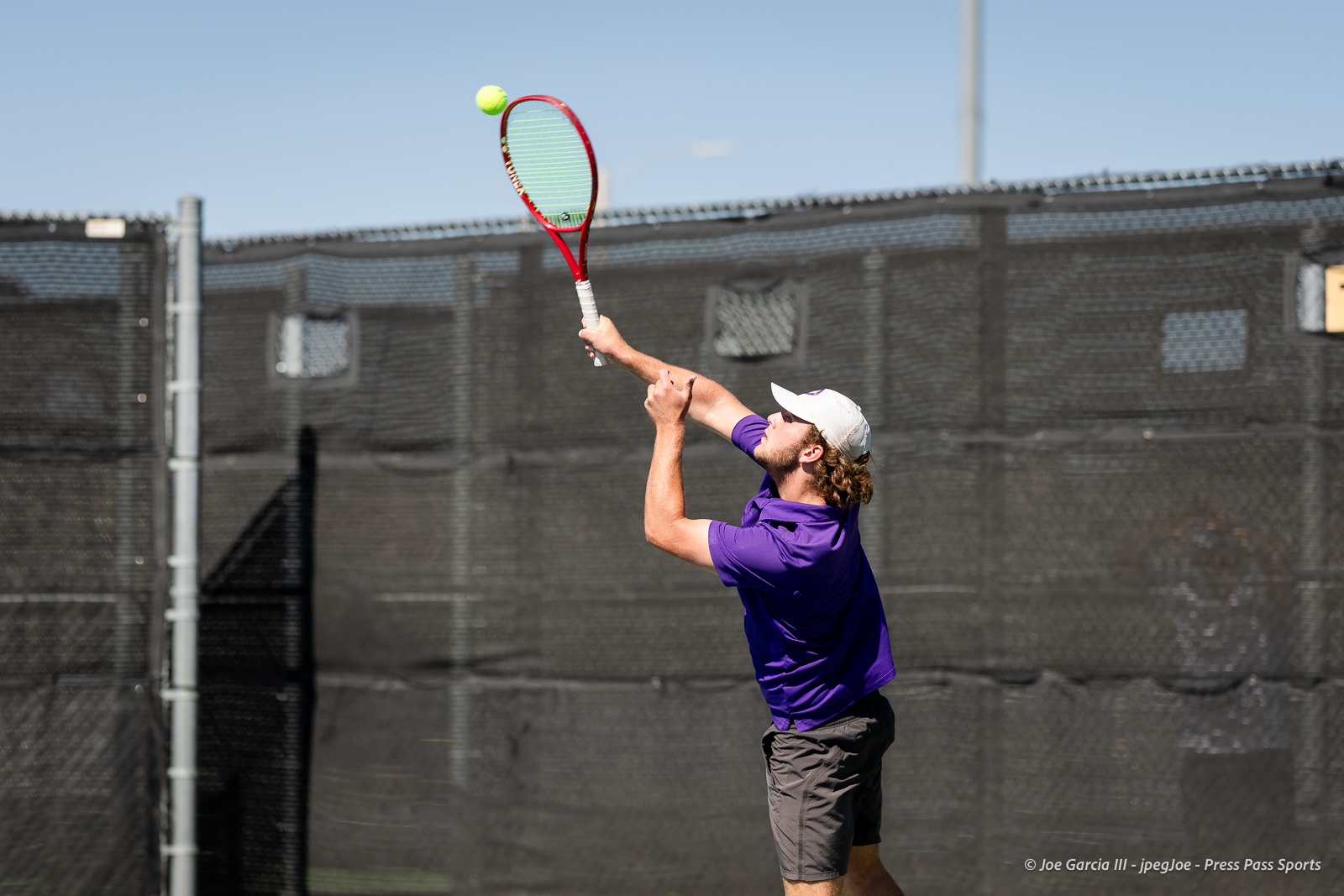 canyon-regional-tennis-75 | Press Pass Sports Tennis player in a purple shirt serves the ball on an outdoor court with a red racket and white cap.