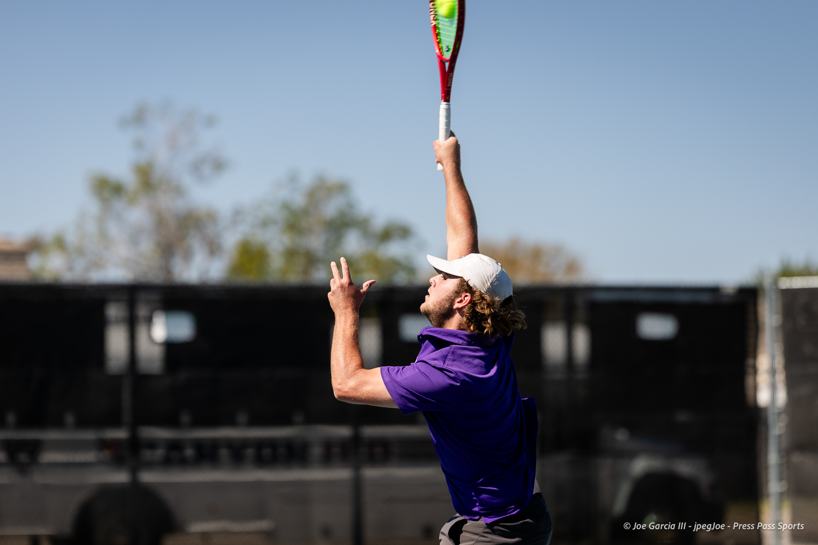 canyon-regional-tennis-77 | Press Pass Sports Male tennis player in a purple shirt and white cap serves the ball with his racket raised high on an outdoor court.