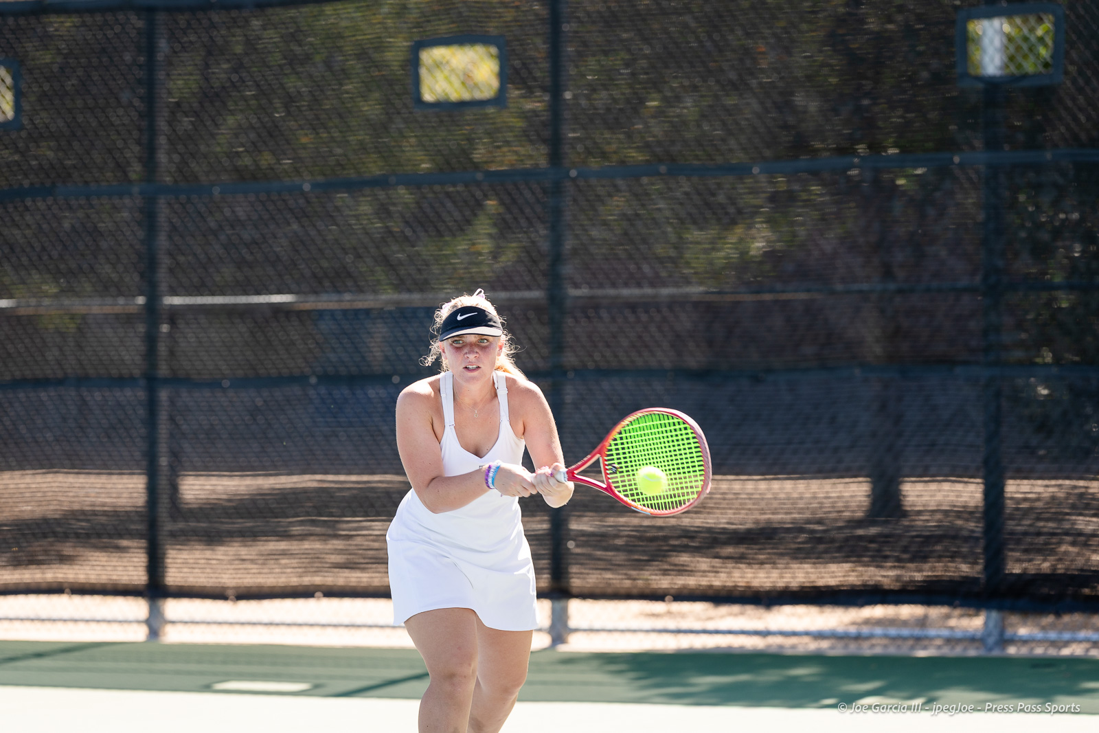 canyon-regional-tennis-80 | Press Pass Sports Female tennis player in a white dress and visor swings at a ball on an outdoor court with a chain-link fence in the background.