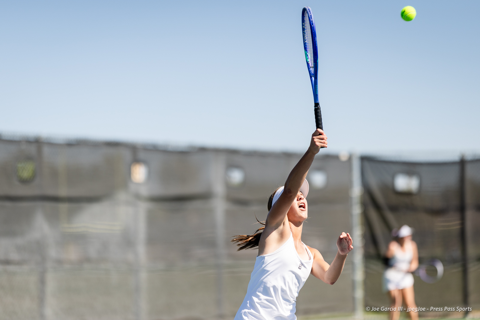 canyon-regional-tennis-84 | Press Pass Sports Female tennis player in a white outfit serves the ball on an outdoor court, racket raised as a bright tennis ball flies overhead.