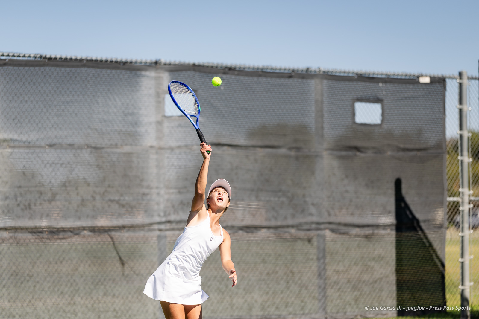 canyon-regional-tennis-85 | Press Pass Sports Female tennis player in a white outfit serving on an outdoor court, blue racket raised as ball sails upward with a chain-link fence backdrop.