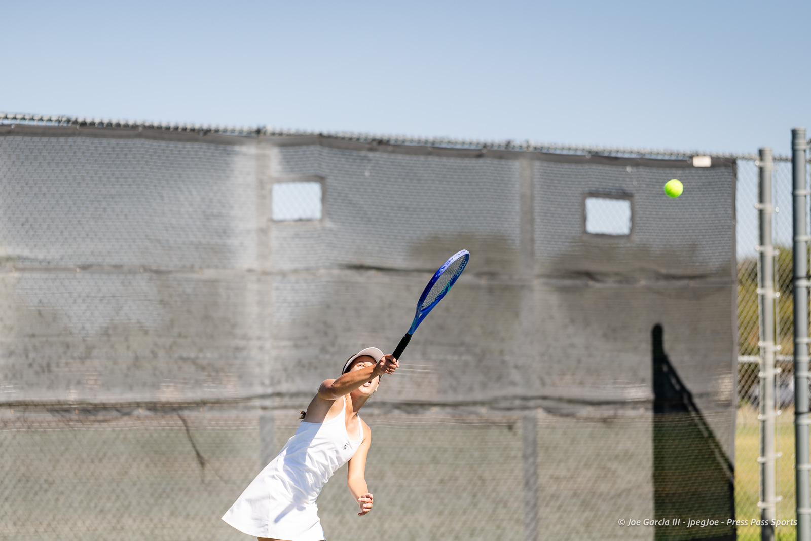canyon-regional-tennis-86 | Press Pass Sports Female tennis player in a white outfit swings a blue racquet, hitting a bright tennis ball on an outdoor court with a chain-link fence backdrop.