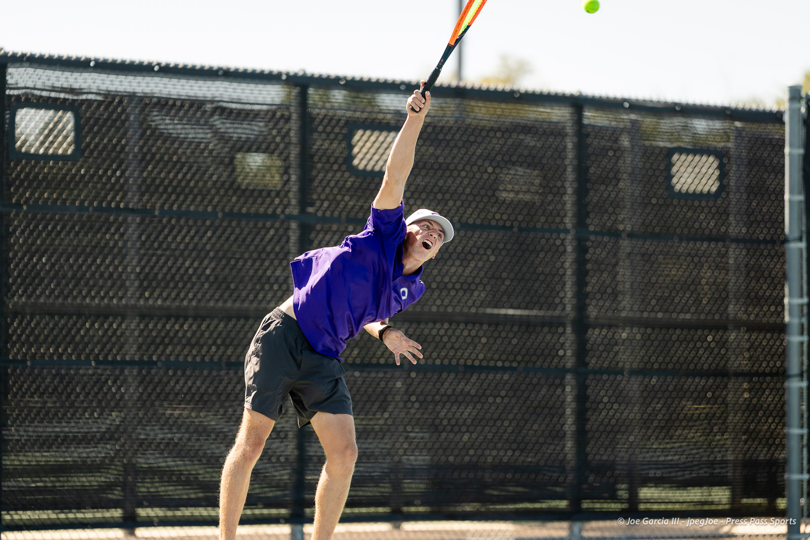 canyon-regional-tennis-88 | Press Pass Sports Male tennis player in a purple shirt leaps to hit a ball with a racket on an outdoor court, chain-link fence in the background.