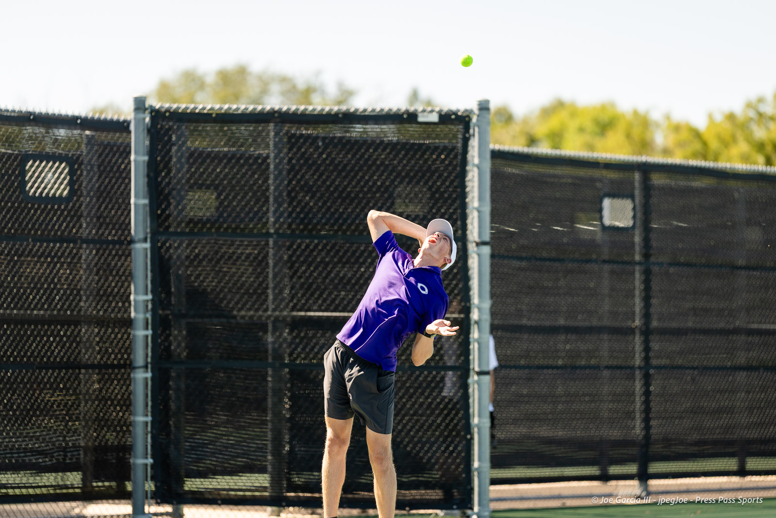 canyon-regional-tennis-89 | Press Pass Sports Male tennis player in a purple shirt serving on an outdoor court with a tall chain-link fence in the background.