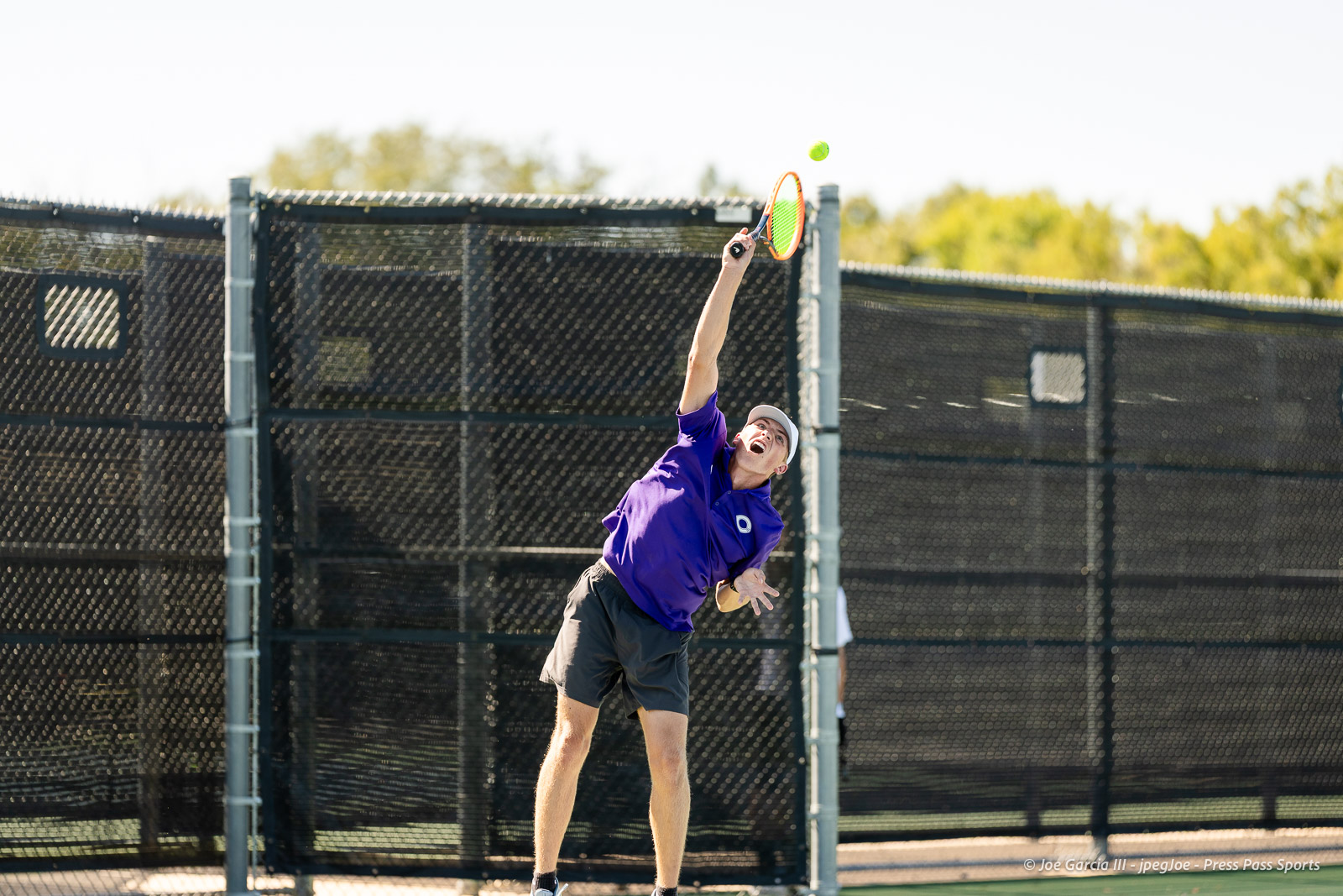 canyon-regional-tennis-90 | Press Pass Sports Tennis player in a purple shirt and cap serves a ball on an outdoor court, orange racket mid-swing.