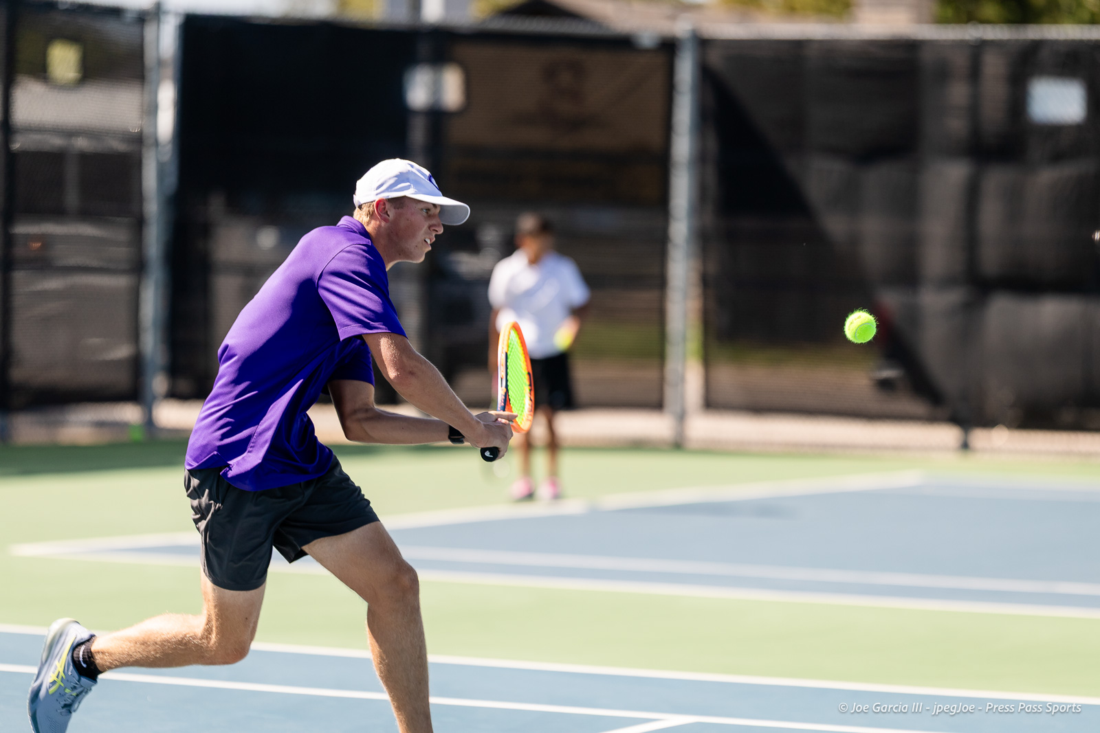 canyon-regional-tennis-91 | Press Pass Sports Male tennis player in a purple shirt and white cap lunges to hit a forehand on an outdoor court; neon-green ball in midair.