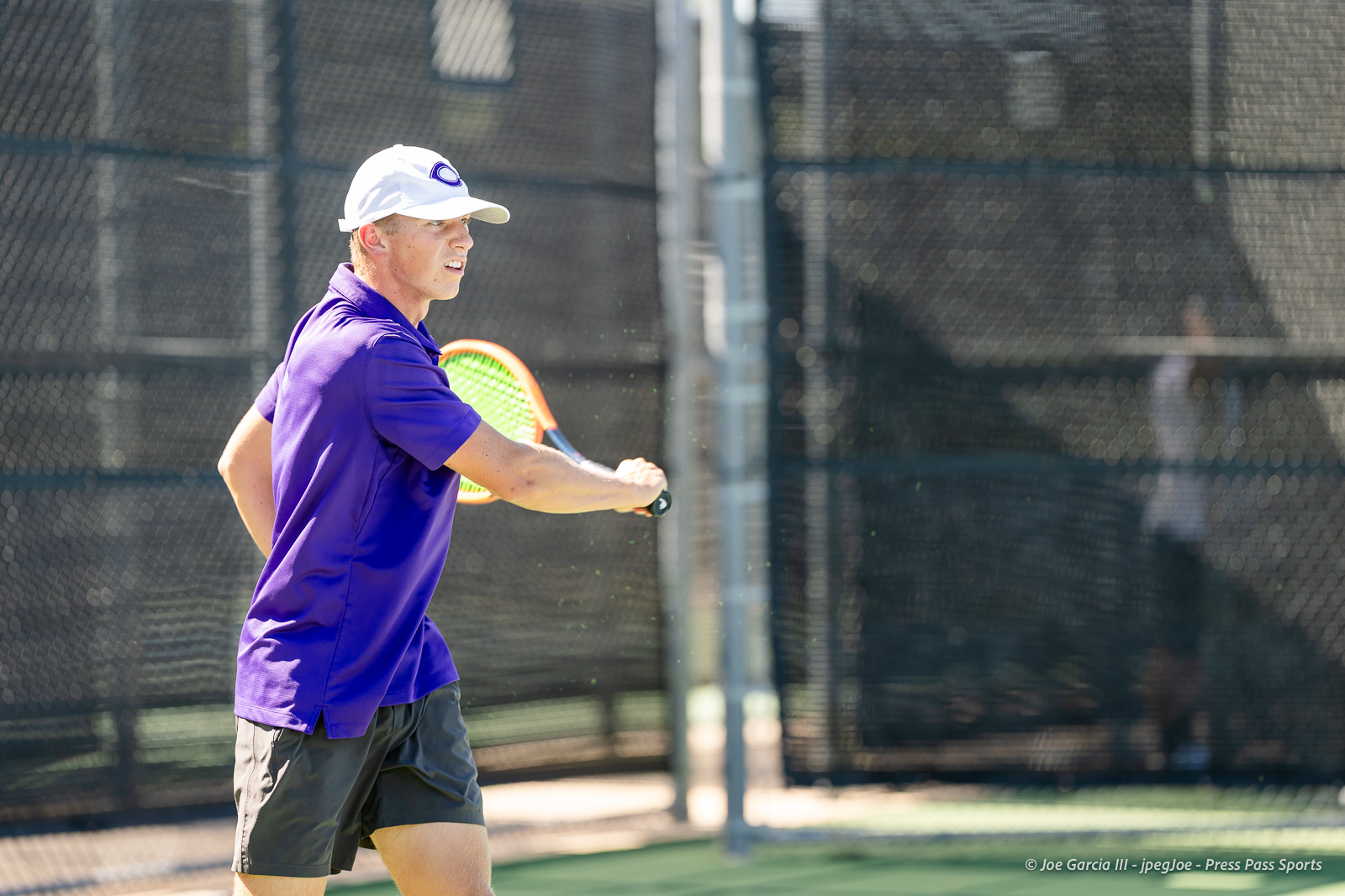 canyon-regional-tennis-95 | Press Pass Sports Tennis player in a purple shirt and white cap swinging a forehand on an outdoor court with a chain-link fence behind him.