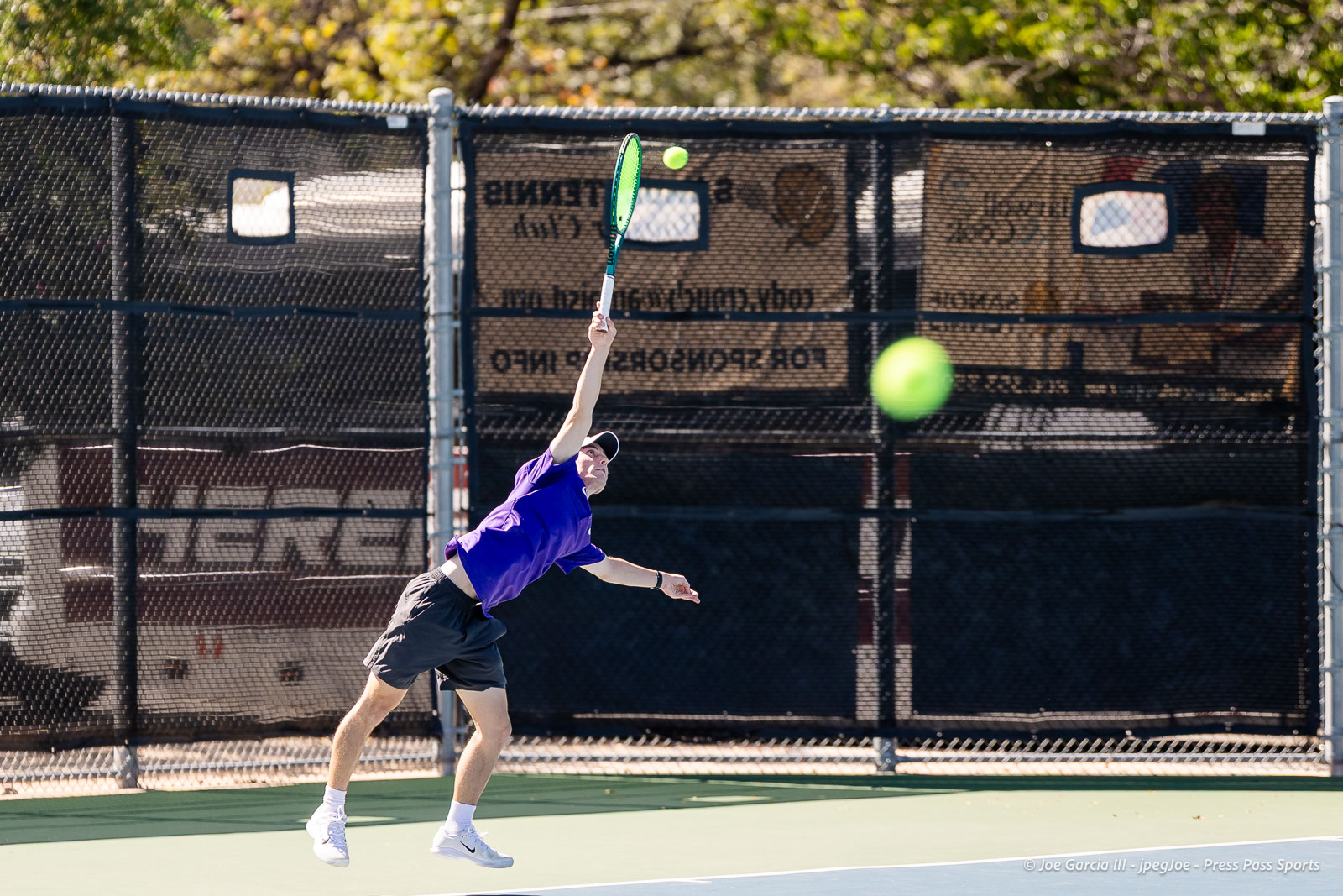 canyon-regional-tennis-98 | Press Pass Sports Tennis player in a purple shirt leaps to serve on an outdoor court, racket raised and ball approaching.