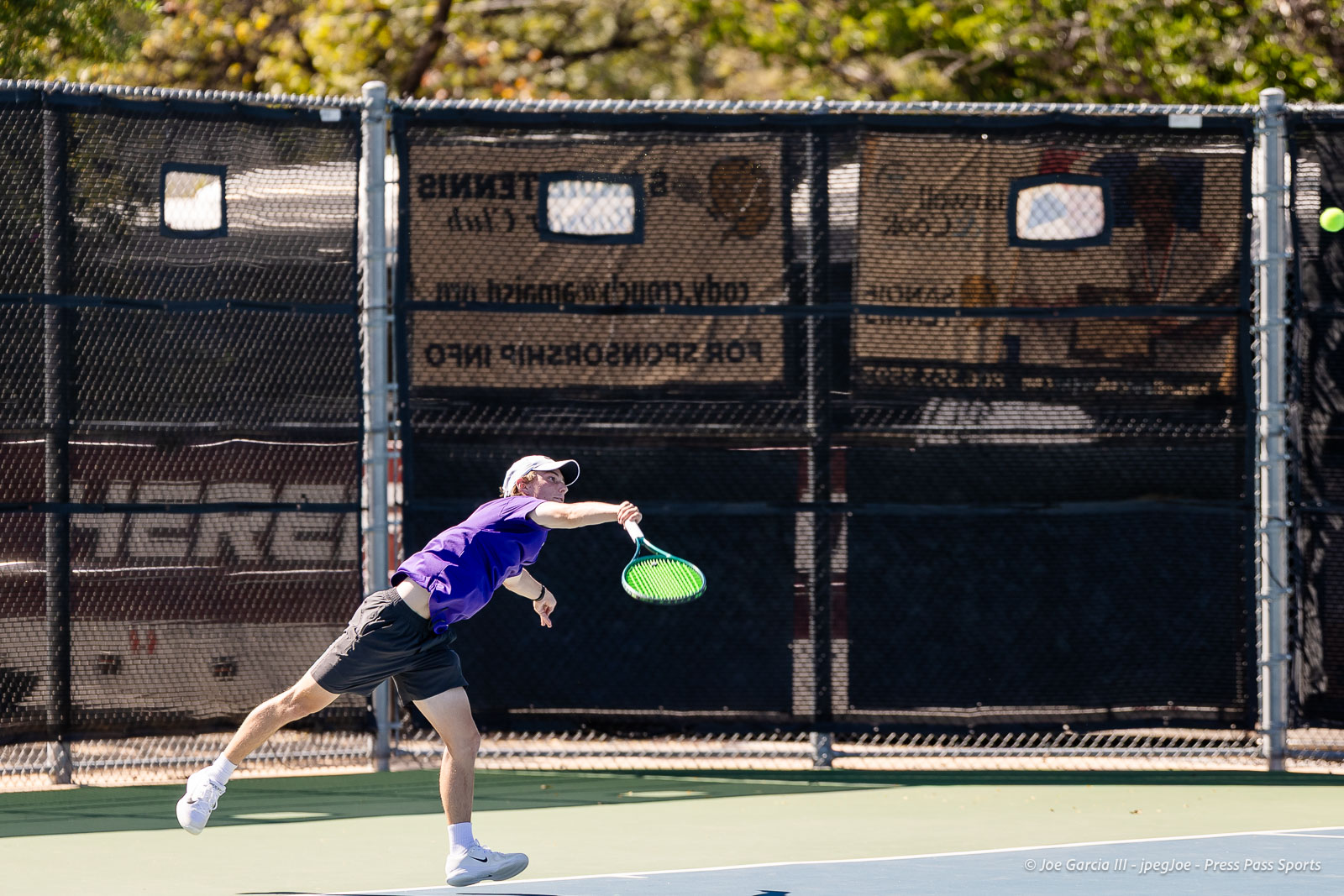 canyon-regional-tennis-99 | Press Pass Sports Tennis player in a purple shirt and white cap lunges to hit a forehand on an outdoor court with a chain-link fence behind.