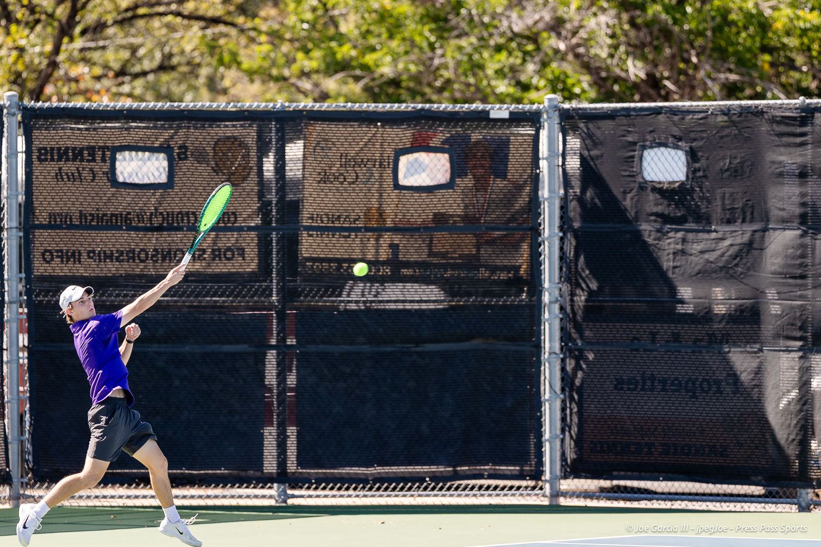 canyon-regional-tennis-100 | Press Pass Sports Tennis player in a purple shirt and white cap swinging a bright green racket to hit a ball on an outdoor court with a black mesh fence behind him