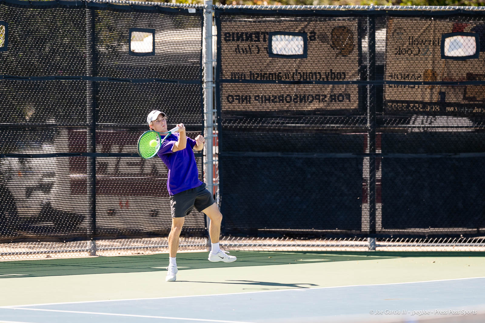 canyon-regional-tennis-101 | Press Pass Sports Tennis player in a purple shirt and white cap mid-swing on an outdoor court with a bright green racket and a chain-link fence behind him.