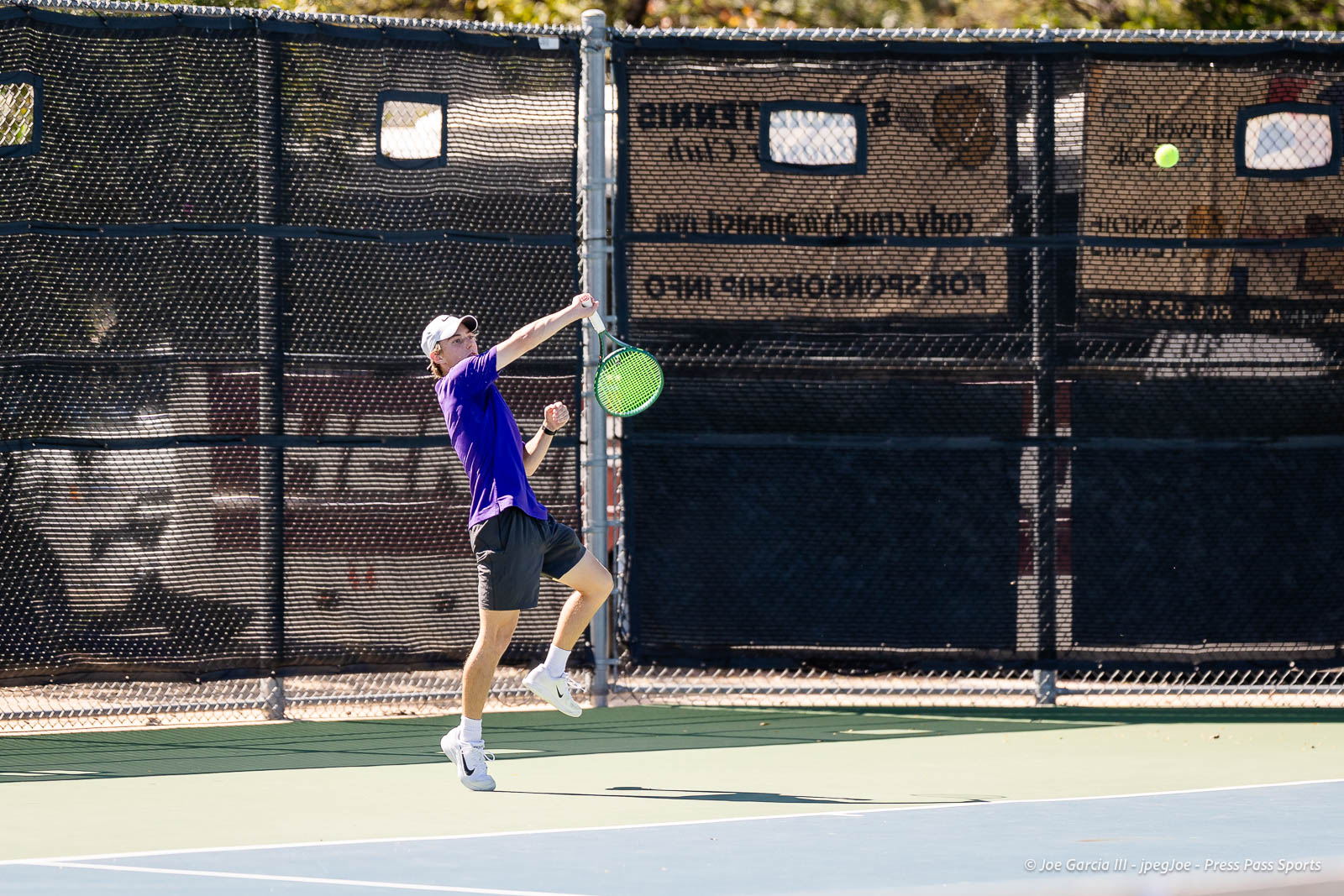 canyon-regional-tennis-102 | Press Pass Sports Male tennis player in a purple shirt and white cap makes a forehand swing on an outdoor court fenced by netting and windscreen behind him.