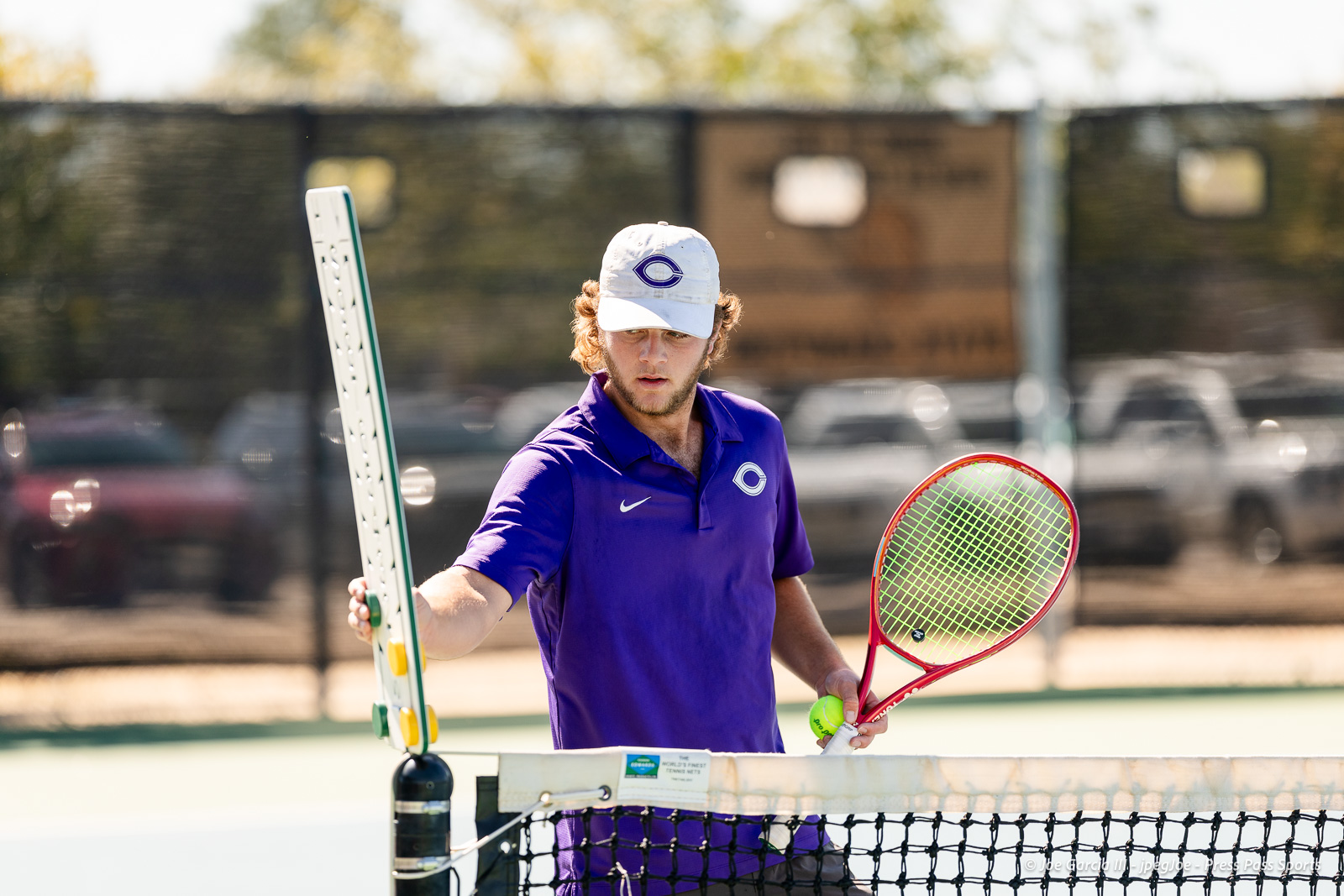 canyon-regional-tennis-103 | Press Pass Sports Male tennis player in a purple polo and white cap at the net, holding a racket in each hand with a ball in one hand on an outdoor court