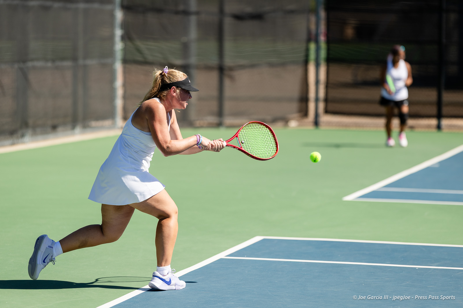 canyon-regional-tennis-104 | Press Pass Sports Female tennis player in a white dress and visor lunges to hit a forehand on a green hard court; opponent blurred in the background.